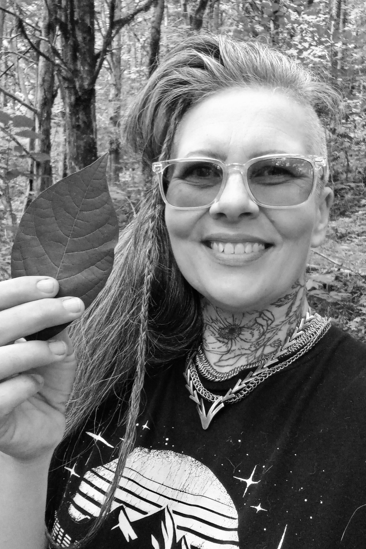 Black and white photo of Melanie Weird, author of Eco-Punk Foraging: How to Use Wild Plants for Food, Medicine, and Crafts. Melanie has long, dark hair and glasses and is wearing a black t-shirt. She is facing the camera and smiling while holding up a leaf.