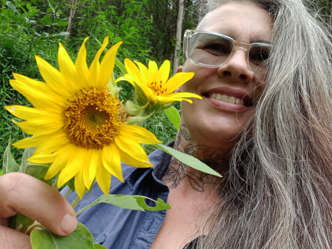 the author of the book Eco-Punk Foraging. She is a middle aged white woman with a warm smile, long salt and pepper hair and glasses. She has a neck tattoo of a rosa rugosa, and is holding up a pair of bright yellow sunflowers towards the camera.